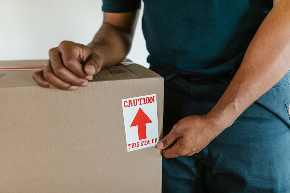 A person wearing a dark green shirt and dark trousers is seen at a house interior, lifting or positioning a large cardboard box with a red and white caution label that reads 'CAUTION THIS SIDE UP' and an upward-pointing arrow. The box appears to be part of a home relocation process, handled by Removal Company Acton, involving careful packing and handling of household items. The setting includes a neutral background with soft lighting, emphasizing the focus on the individual’s hands and the box, indicating careful loading or unpacking within a moving or furniture transport context, aligned with local removals services and packing procedures.